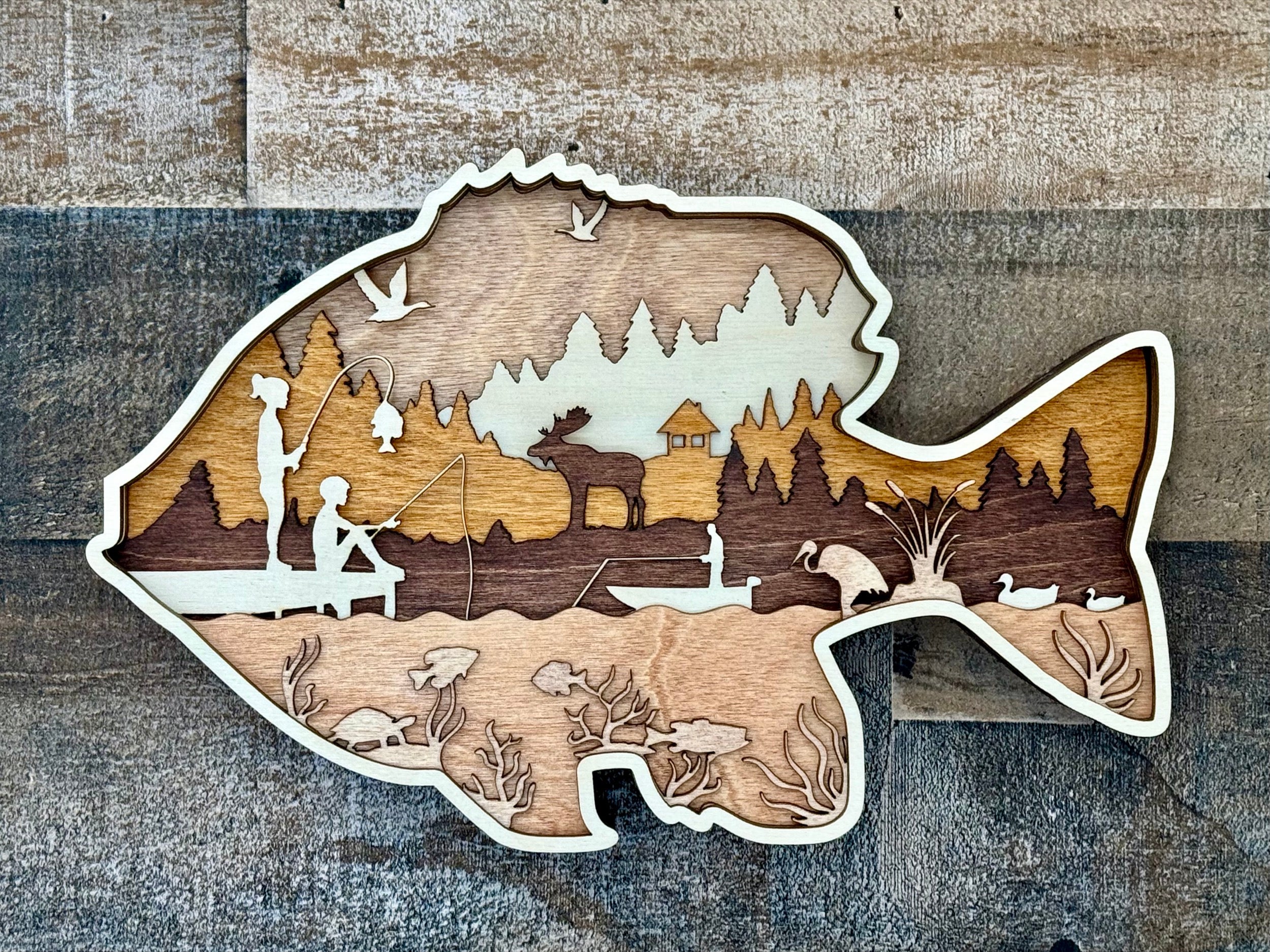 Wooden cutout of a fish with a landscape inside, featuring people fishing and nature scenes.